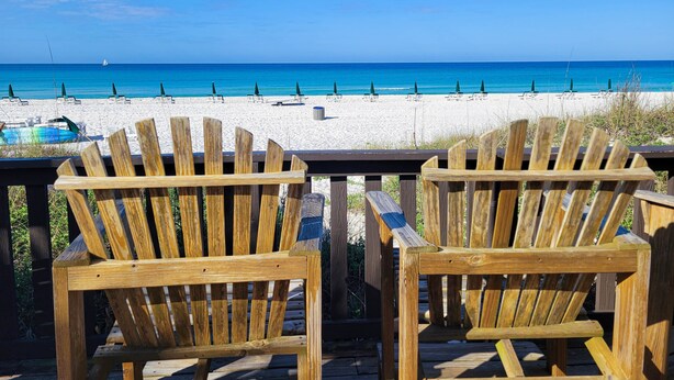 Twee houten Adirondack-stoelen gericht op een strand met een heldere lucht.