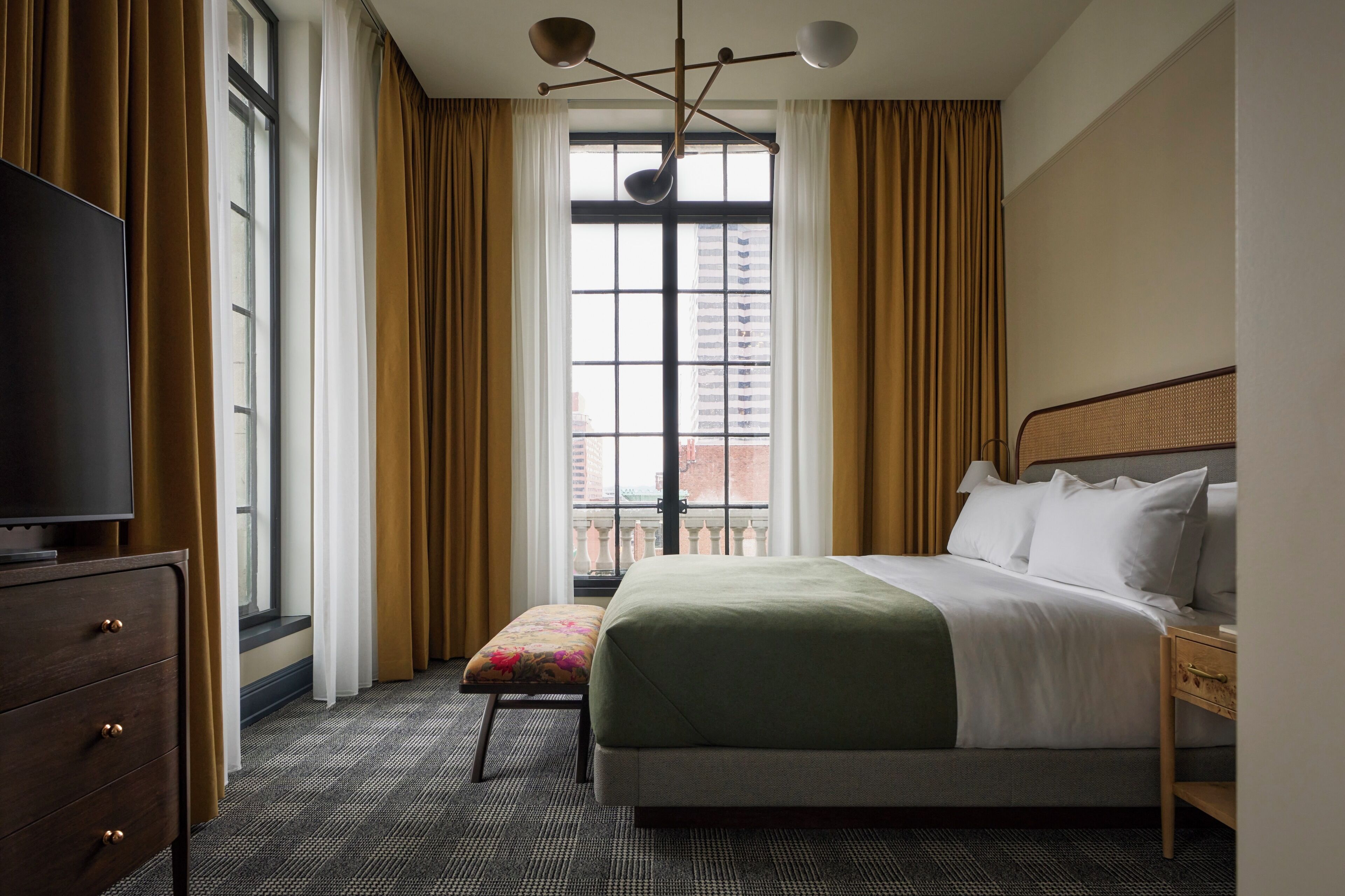 A hotel room with a bed, a television, a window with curtains, and a view of a building.