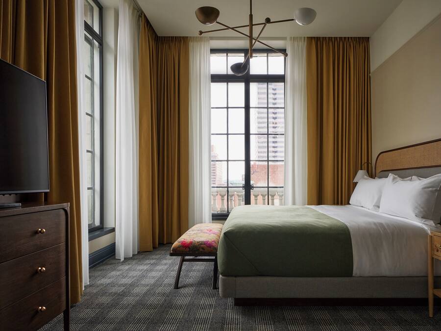 A hotel room with a bed, a television, a window with curtains, and a view of a building.