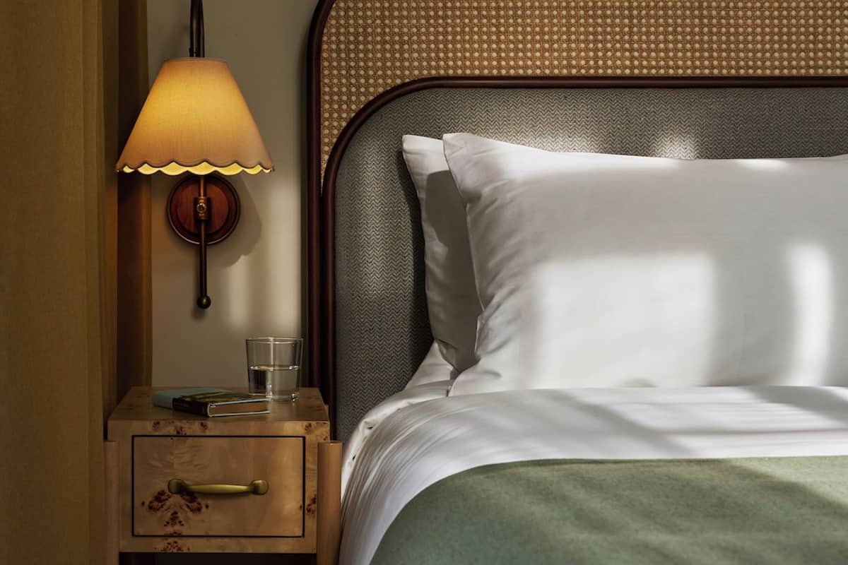 A neatly made bed with a green blanket, a bedside lamp, and a small wooden nightstand with a drawer.
