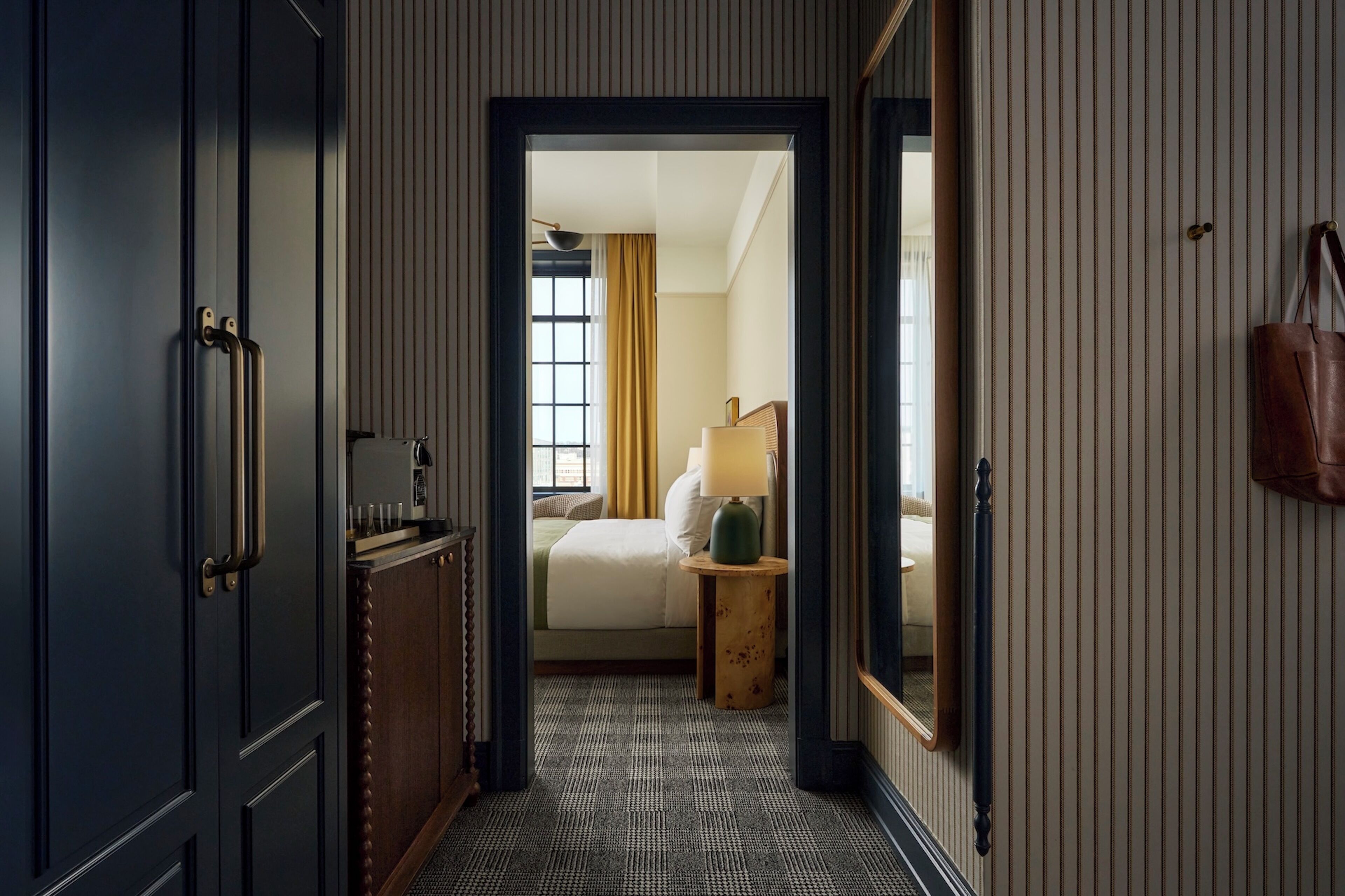 A hotel hallway with a bed, a lamp, and a handbag on the wall.