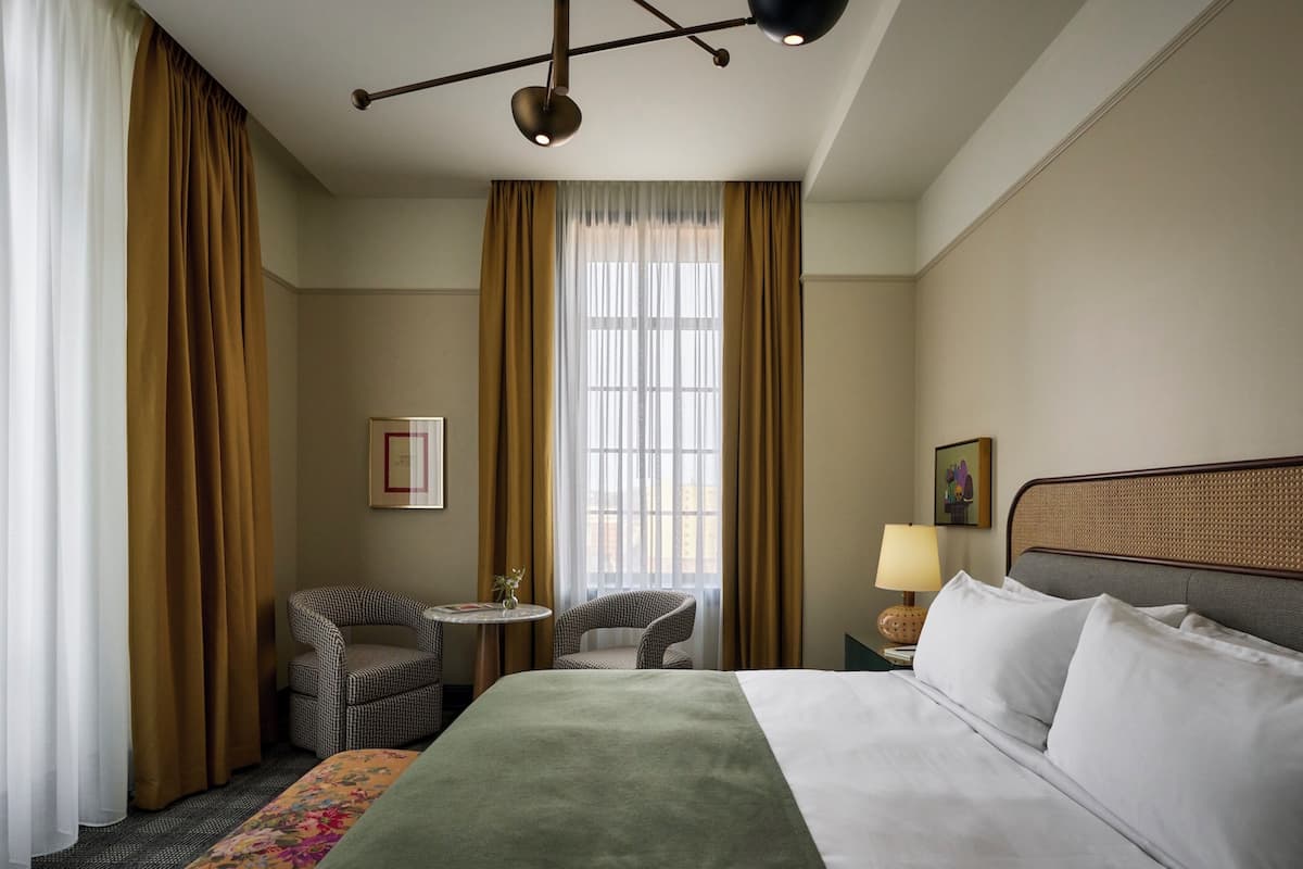 A hotel room with a bed, two chairs, a small table, a lamp, and a window with curtains.