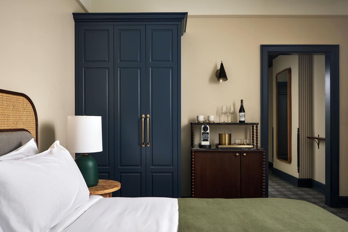 A hotel room with a bed, a wardrobe, a bedside table with a lamp, a small cabinet with a radio, and a mirror in the corner.