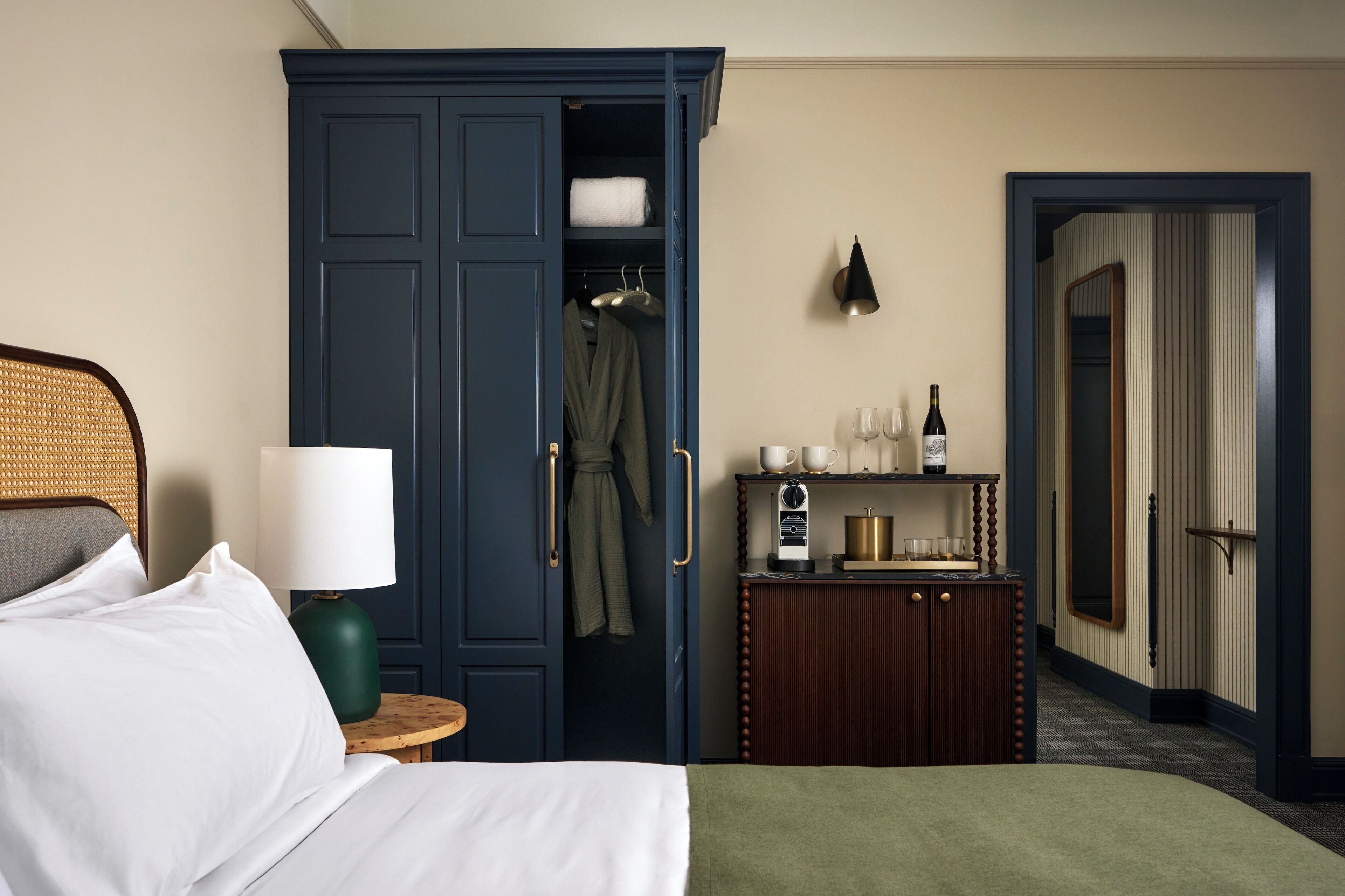 Habitación de hotel con cama, mesita de noche con lámpara, armario, pequeño mueble con radio y espejo.