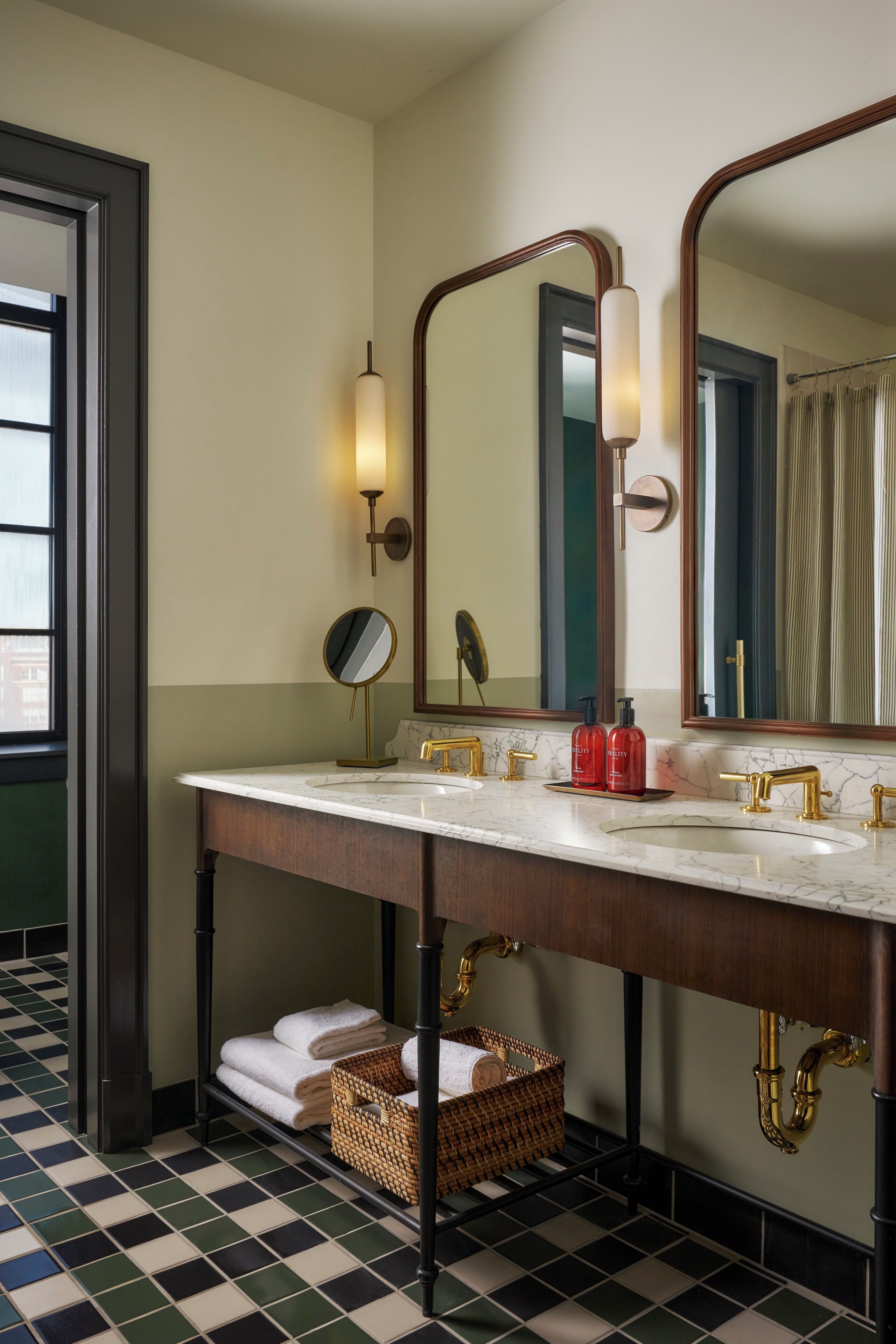 A bathroom with a double sink, two mirrors, a towel rack, and a basket.