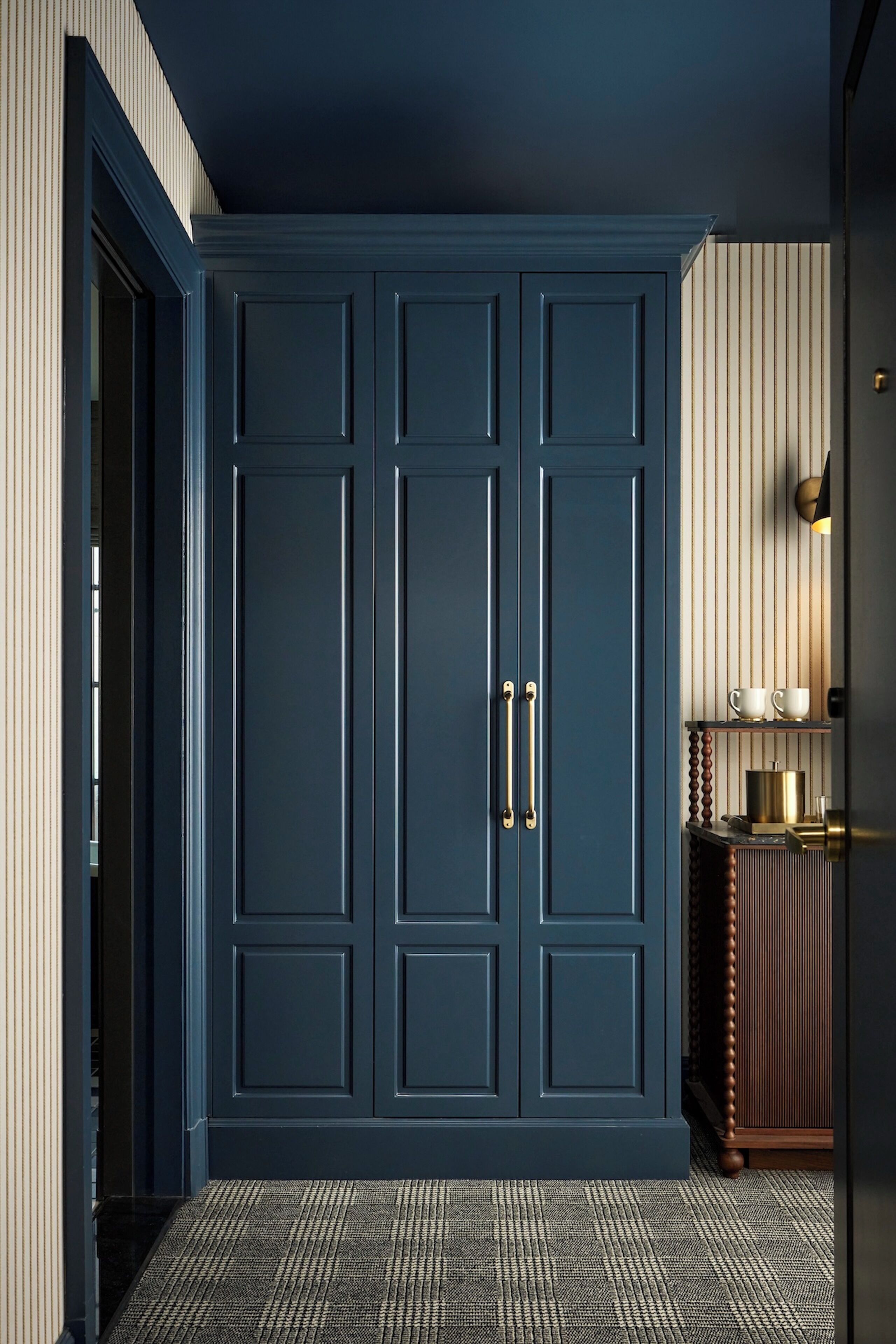Una habitación con un armario azul oscuro grande, una alfombra estampada y una mesita de madera con una lámpara.