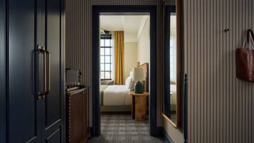 A hallway leading to a bedroom with a bed, bedside table, lamp, and a hanging bag.