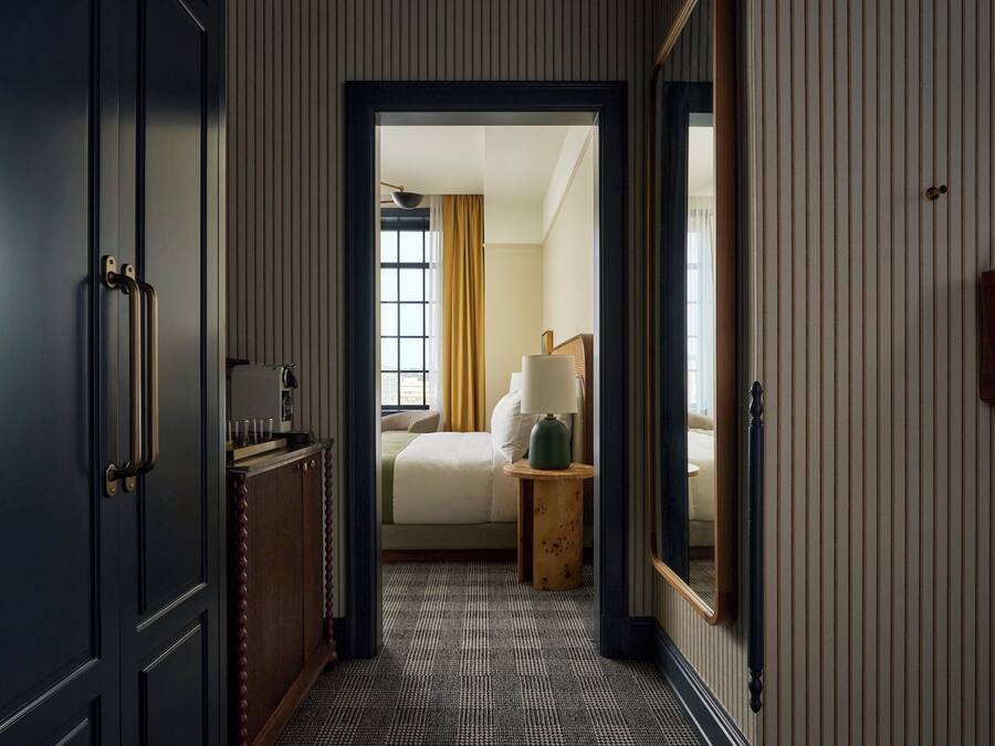 A hallway leading to a bedroom with a bed, bedside table, lamp, and a hanging bag.