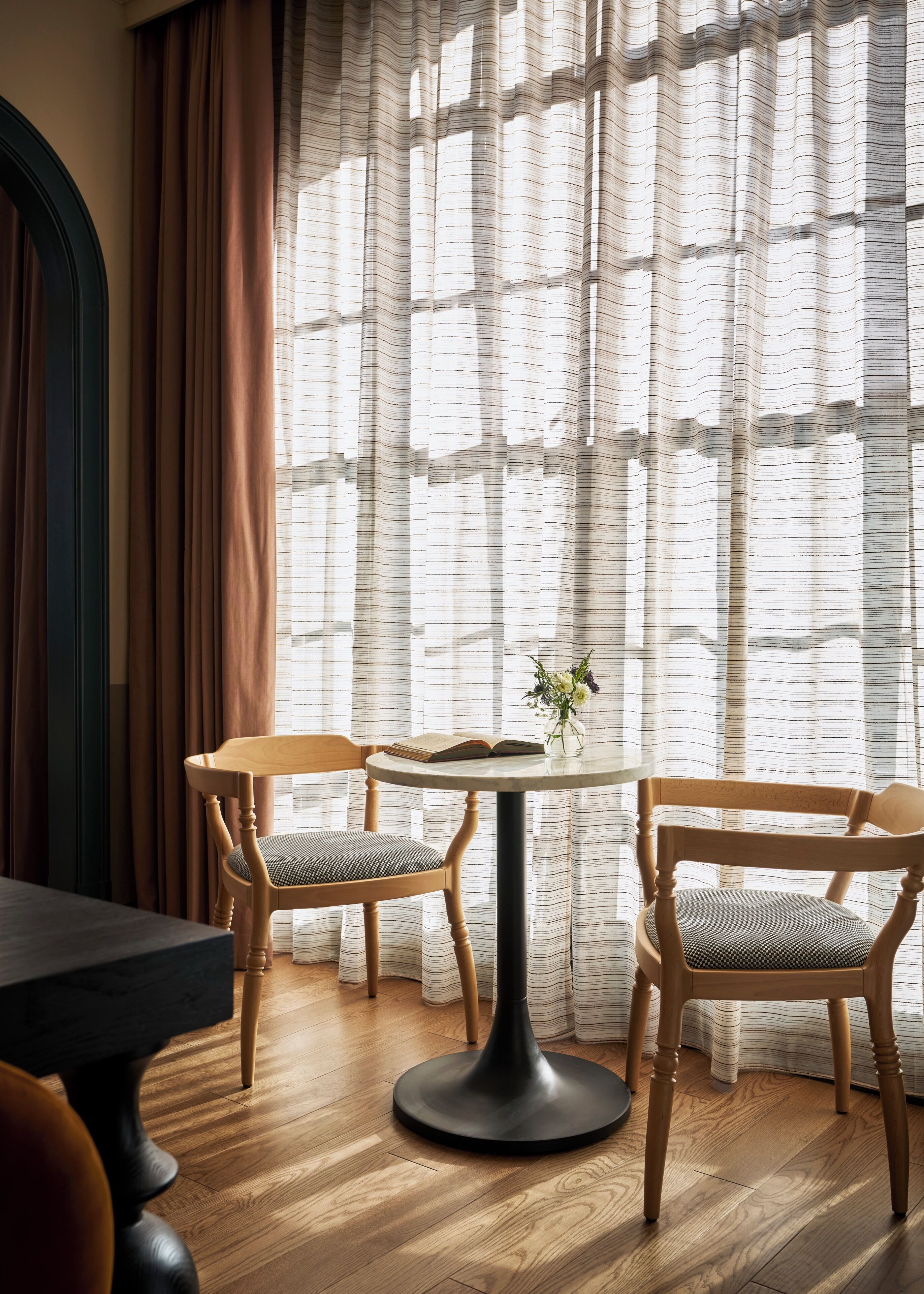 A dining area with a round table, two wooden chairs, a vase with flowers, and a window with sheer curtains.