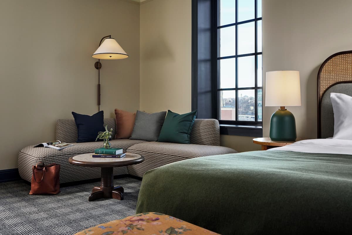 A hotel room with a sofa, a round table, a lamp, a bed, and a window with a view.