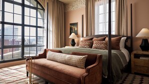 A hotel room with a large window, a bed with multiple pillows, a side table, and a patterned rug.