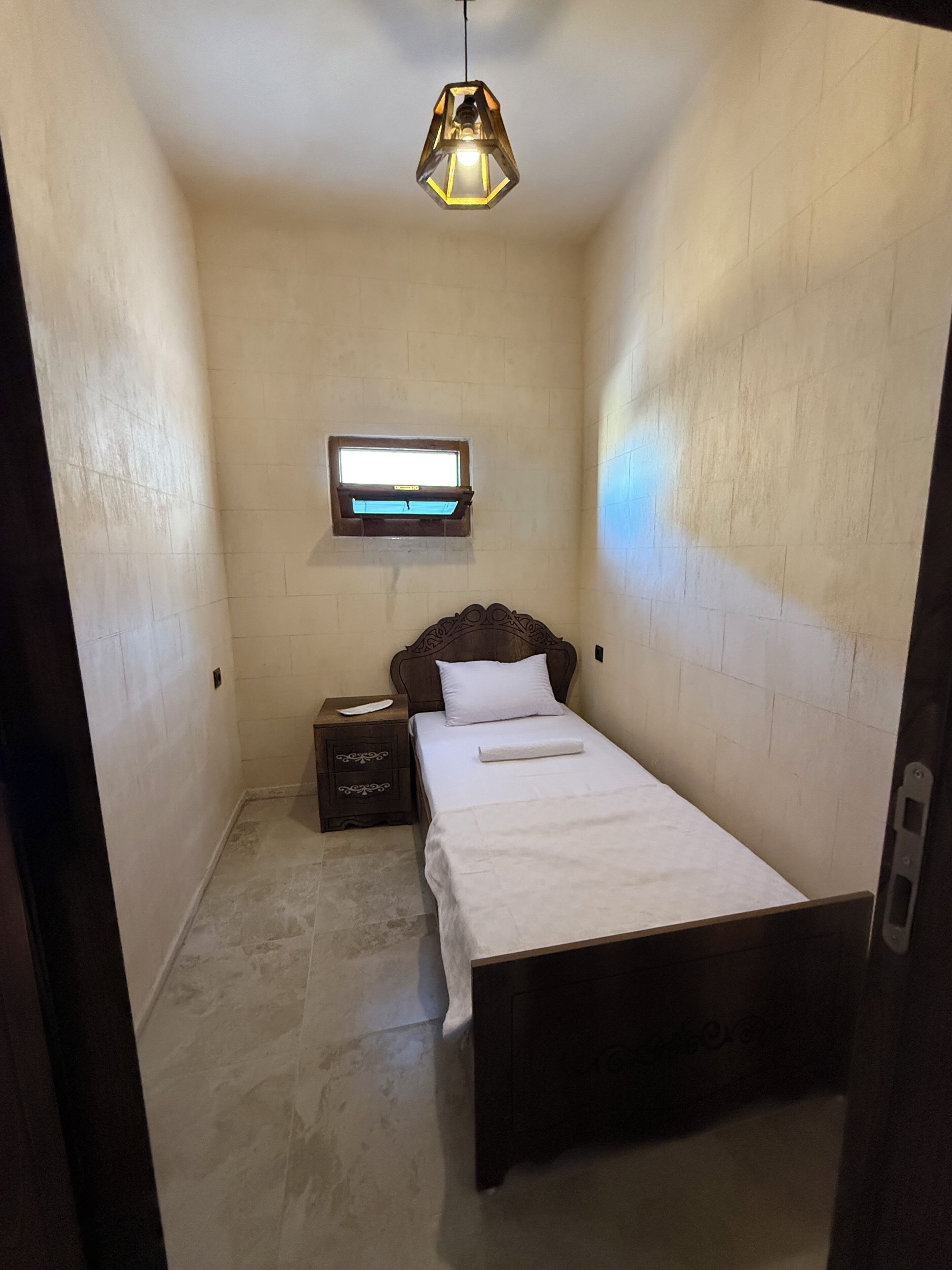Una habitación pequeña y estrecha con una cama individual, una mesita de noche, un televisor instalado en la pared y una lámpara colgante.