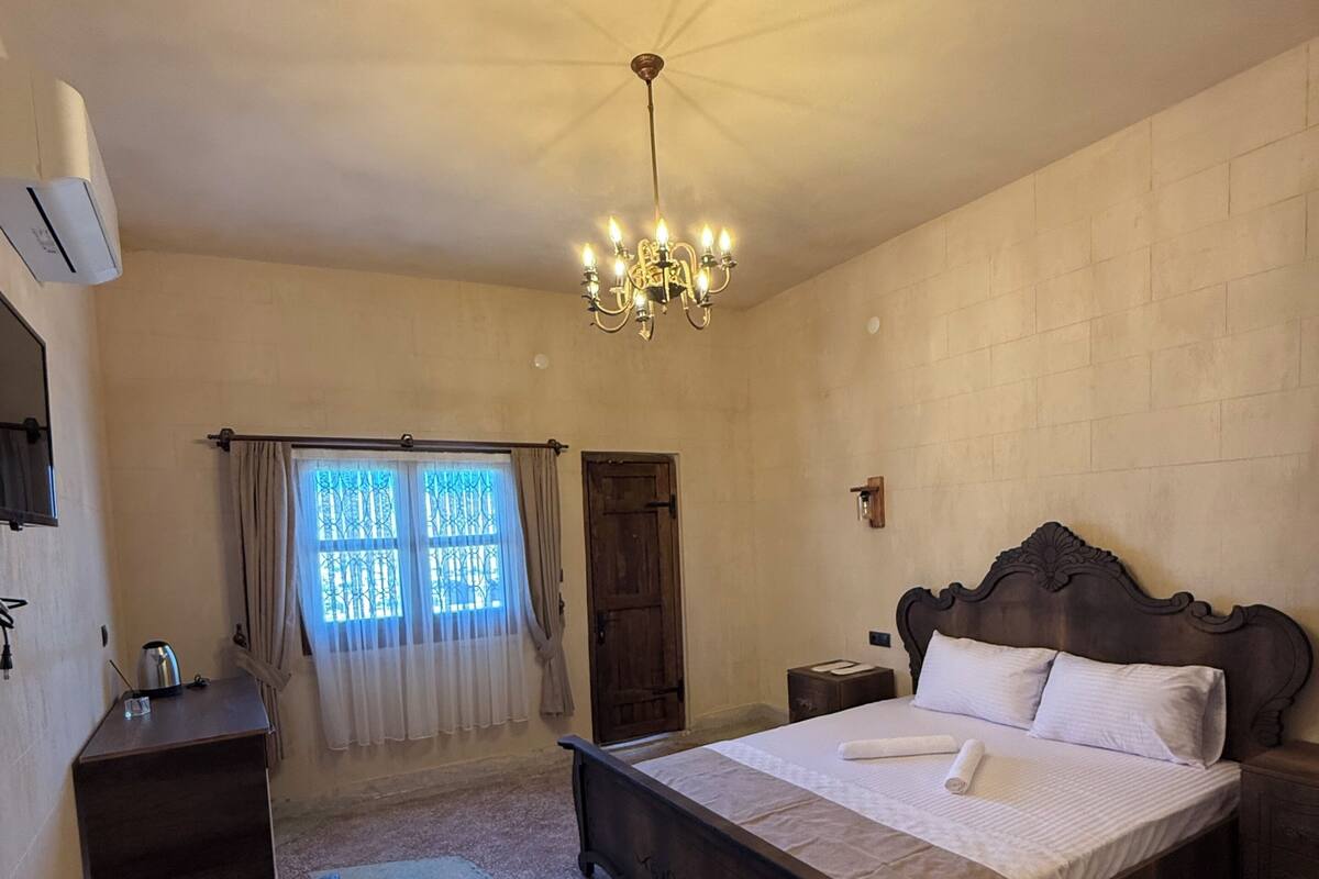 A bedroom with a large bed, a bedside table, a dresser, a chandelier, and a window with curtains.