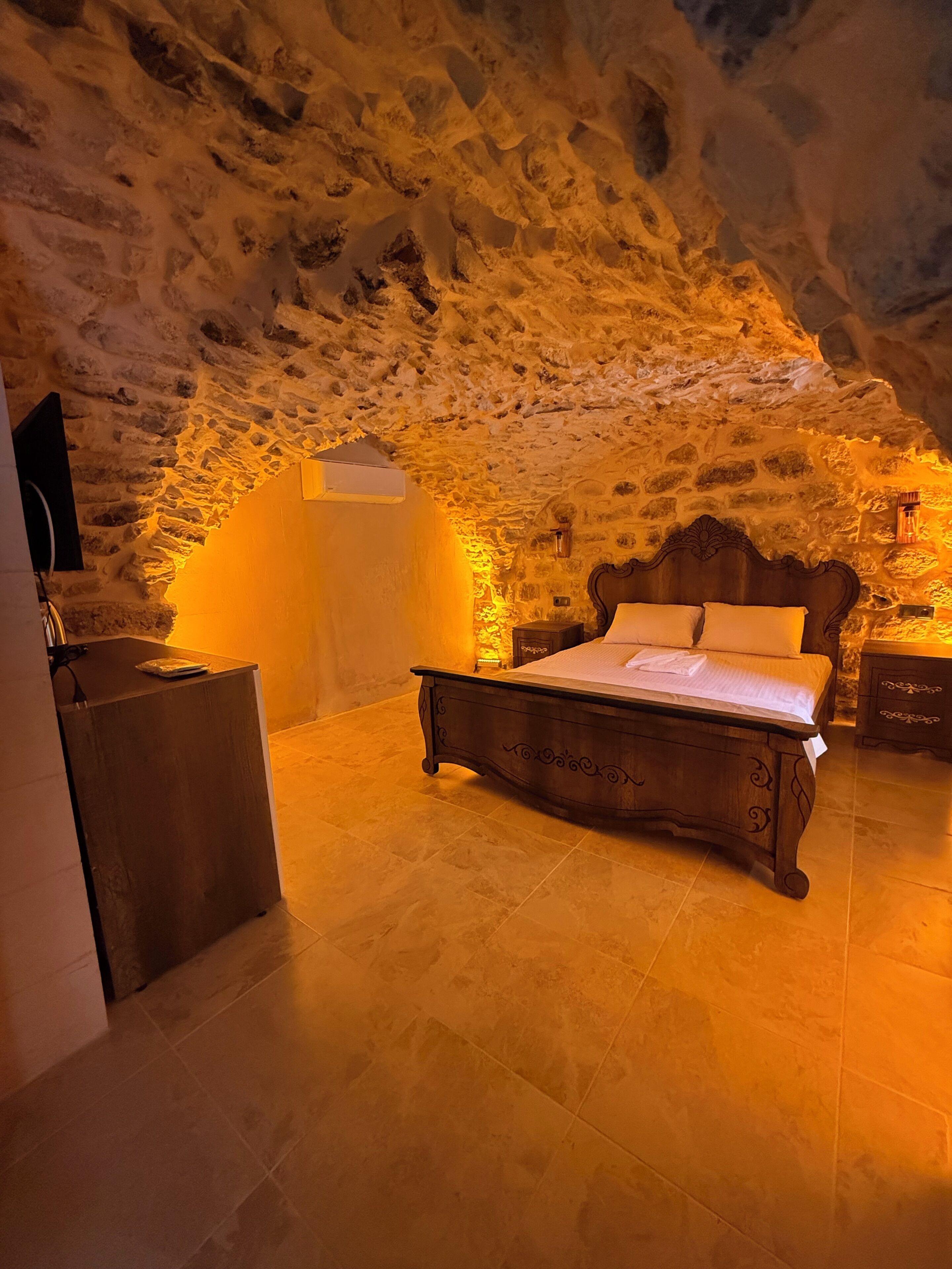 A bedroom with a stone wall, a wooden bed, a bedside table, and a small cabinet.