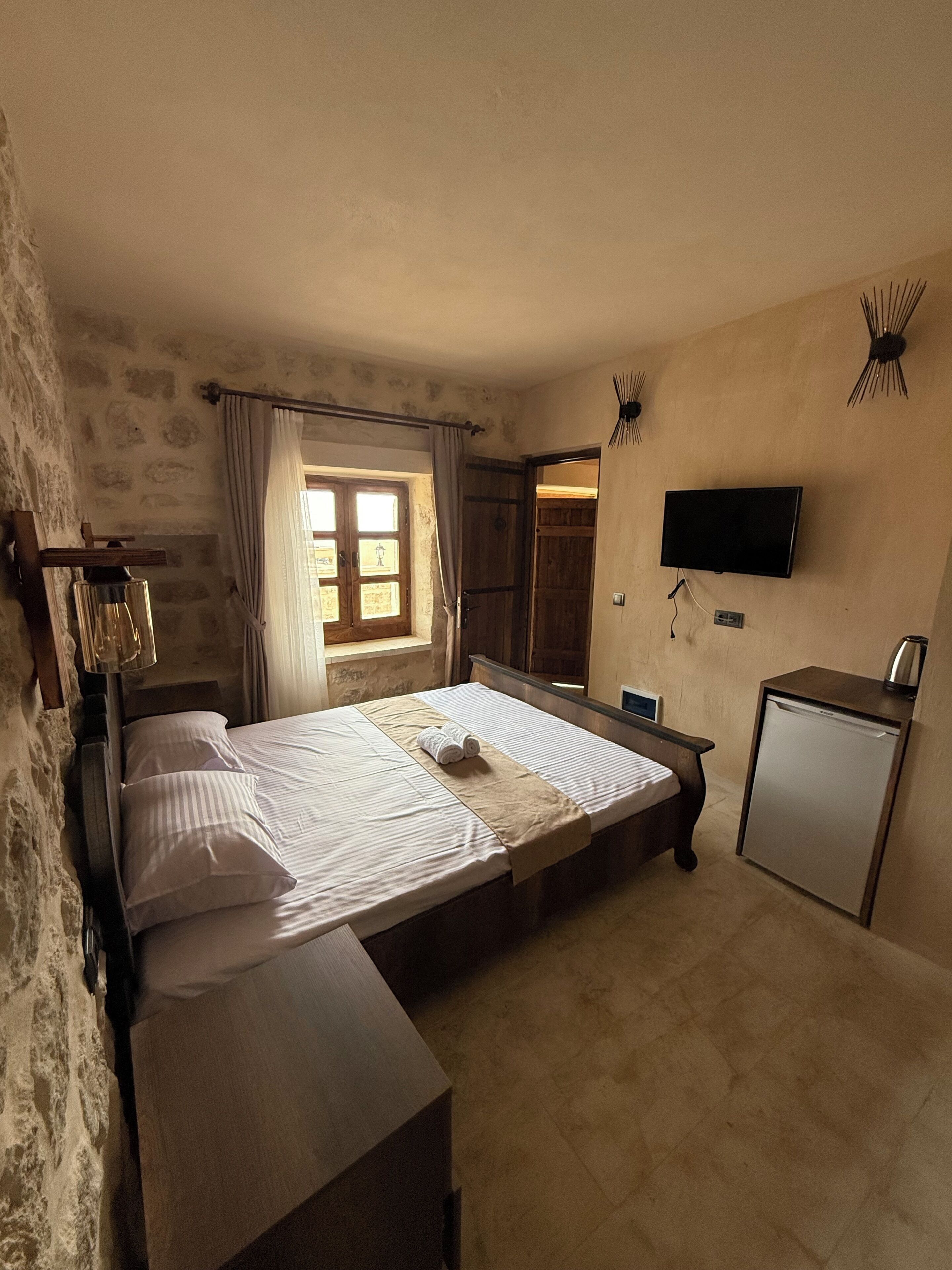 A bedroom with a bed, a small desk, a television, and a window with curtains.