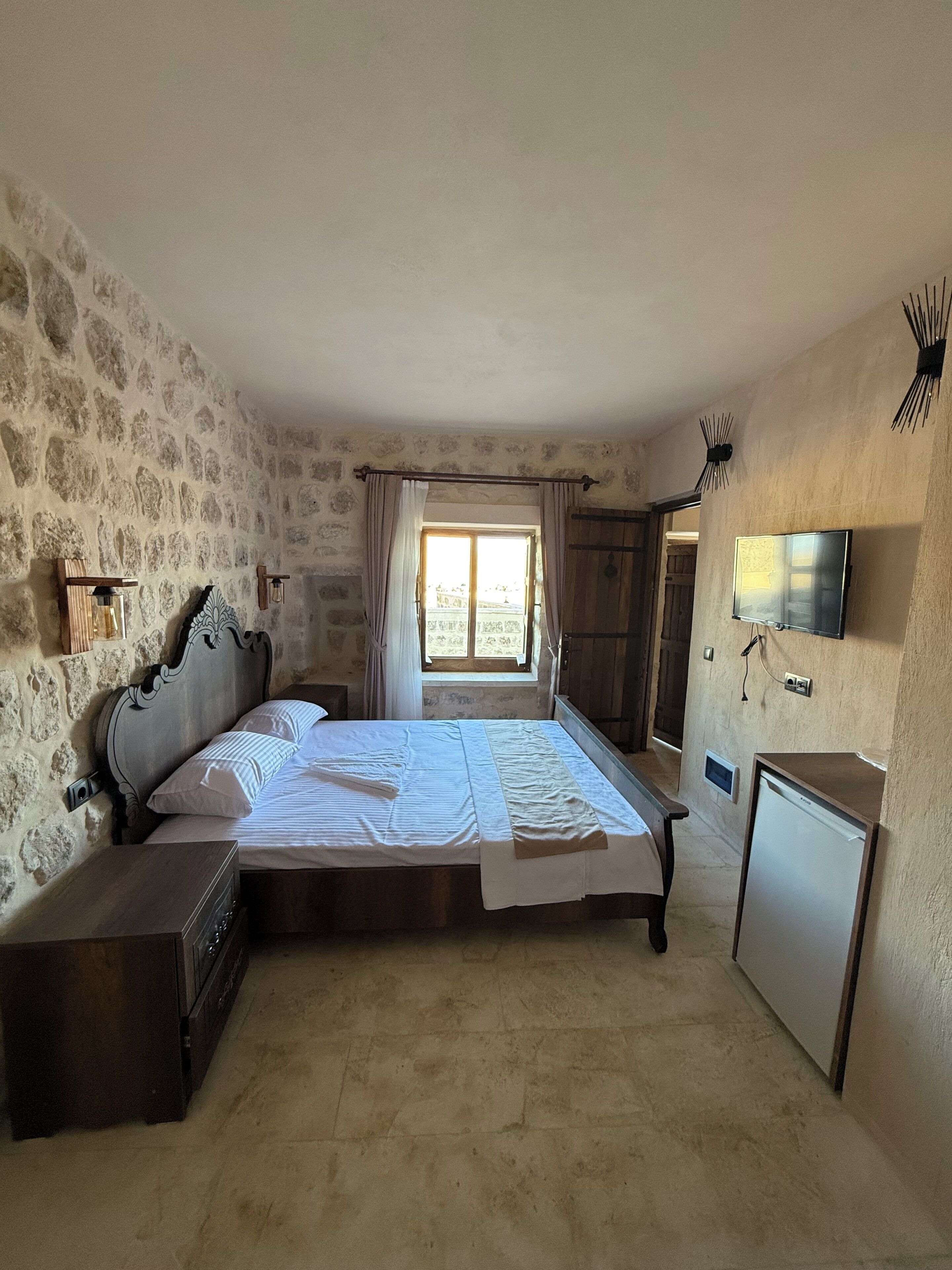 Un dormitorio con pared de piedra, una cama con ropa blanca, un escritorio pequeño, un televisor y una ventana con cortinas.