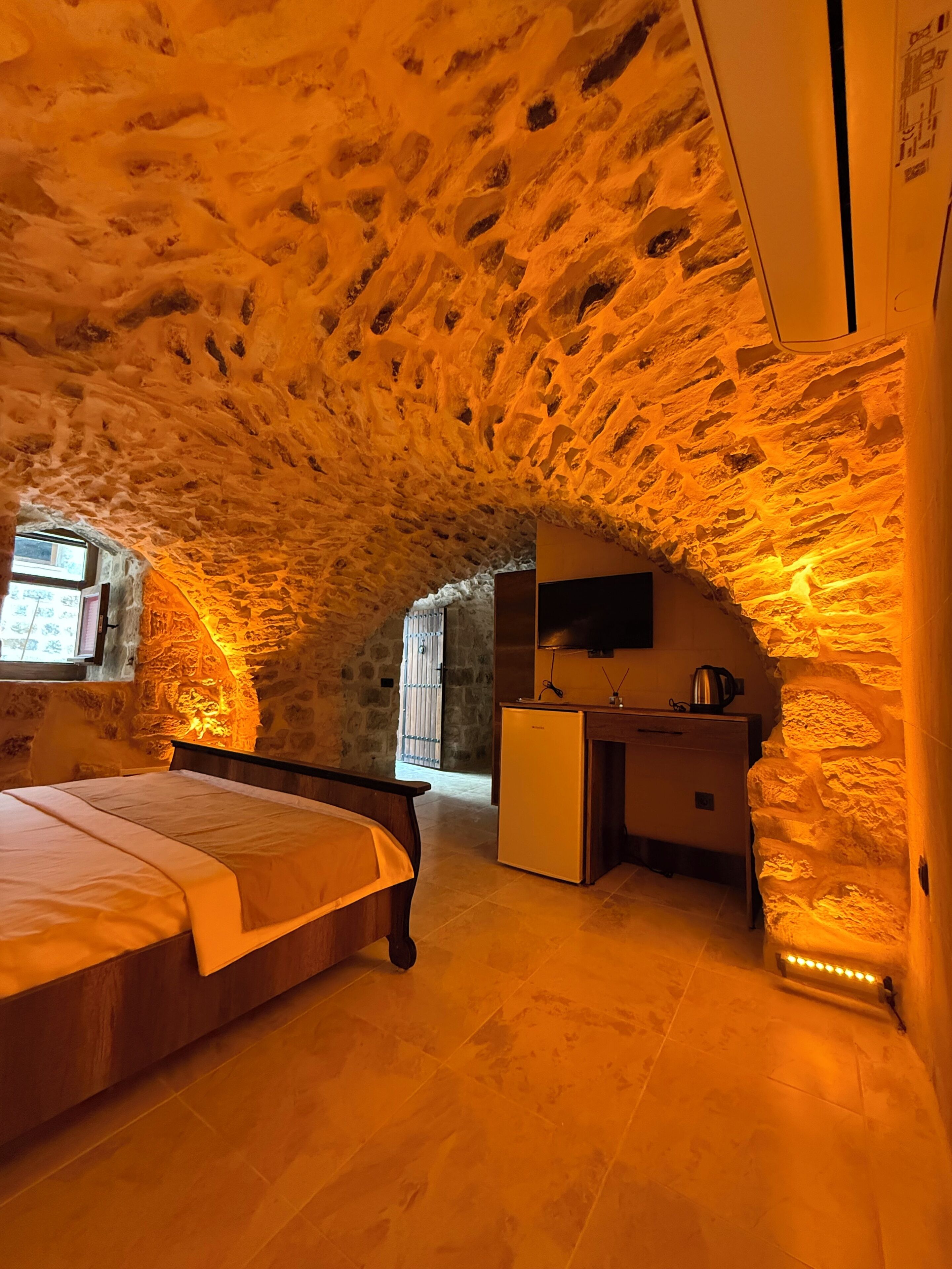 Habitación con paredes de piedra, una cama, un escritorio y un refrigerador.