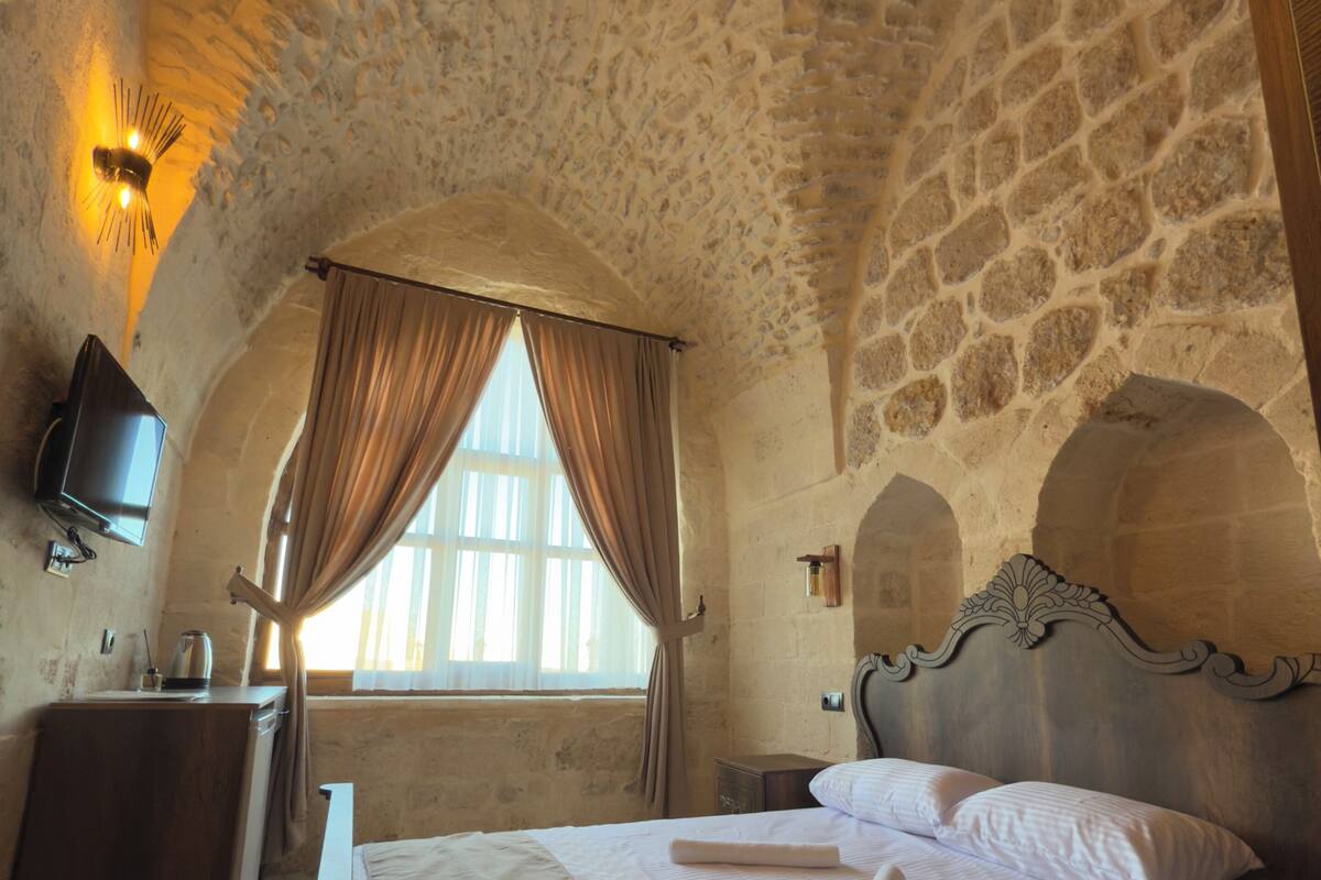 A room with a stone ceiling, a bed with white linens, a television on the wall, and a window with curtains.