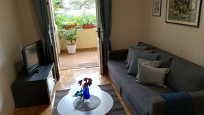 Sala de estar com sofá cinza, mesa de centro com um vaso, televisão em um suporte e vista para um jardim através de uma porta aberta.