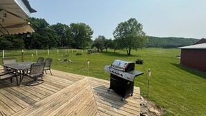 A wooden deck with a grill, outdoor furniture, and a view of a grassy field and trees. - Experience a Farm getaway! (Bagley)