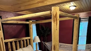 A wooden four-poster bed with a checkered bedspread, a nightstand with a lamp, and a hammock in a room with wooden floors and a red accent wall. - Experience a Farm getaway! (Bagley)