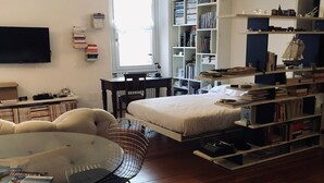 A living room with a sofa, a round glass table, a chair, a bed, a bookshelf, and a TV.