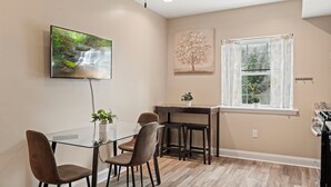 Un comedor con mesa de vidrio, dos sillas, un televisor montado en la pared, un cuadro, una ventana con cortinas y una zona de cocina con estufa.