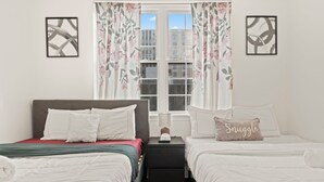 Habitación doble con dos camas, una mesita de noche, una ventana con cortinas florales y obras de arte enmarcadas en las paredes.