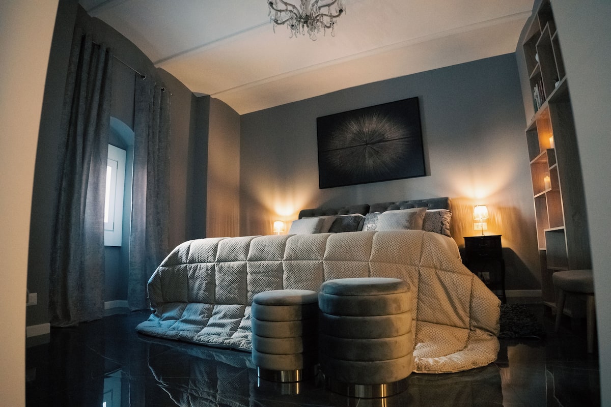 A bedroom with a large bed, two side tables, a chandelier, and a painting on the wall.