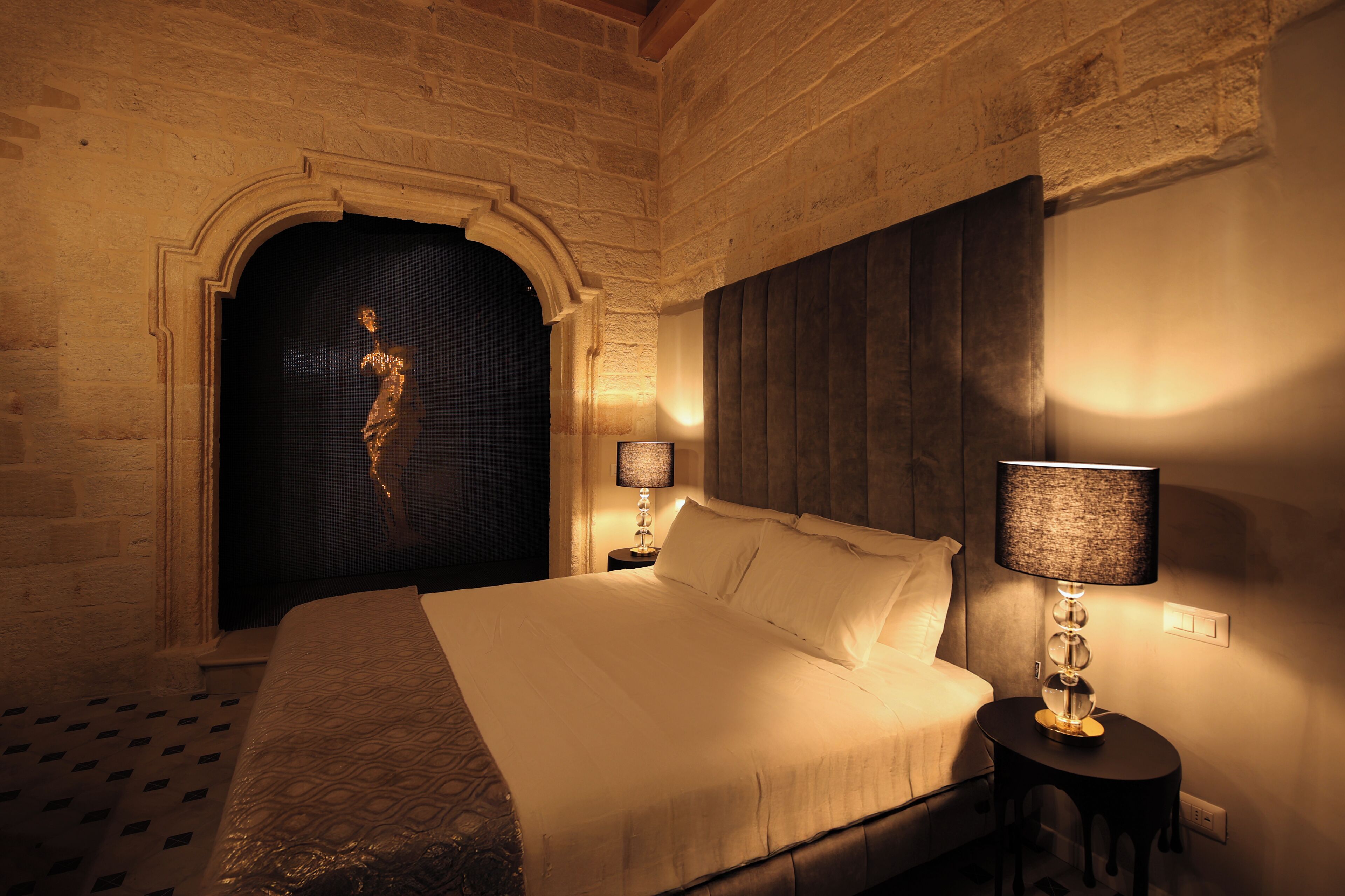 Une chambre d’hôtel avec un grand lit, une table de chevet avec une lampe, une porte cintrée sombre et un mur en briques.