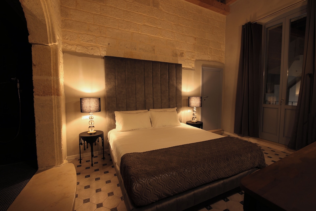 Un dormitorio con una cama grande, dos mesitas de noche, un cabecero y una puerta con cortinas.