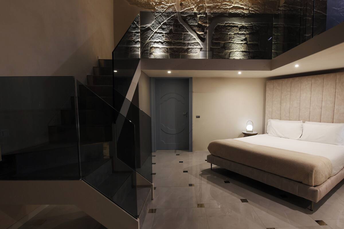 Une chambre moderne avec un grand lit, un escalier en verre et un mur en pierre.