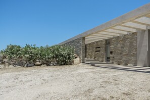 A modern building with a stone facade, a covered entrance, and a gravel courtyard. - Aetoula: Charming Stone Cave House with Sunset Views in Mylopotas (Ίος)