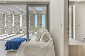 A modern bedroom with a bed, a blue towel, and a view of stone pillars through a glass door. - Aetoula: Charming Stone Cave House with Sunset Views in Mylopotas (Ίος)