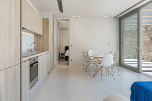 A modern kitchen with a built-in oven, a dining area with a table and chairs, and a large sliding glass door leading to an outdoor area. - Aetoula: Charming Stone Cave House with Sunset Views in Mylopotas (Ίος)