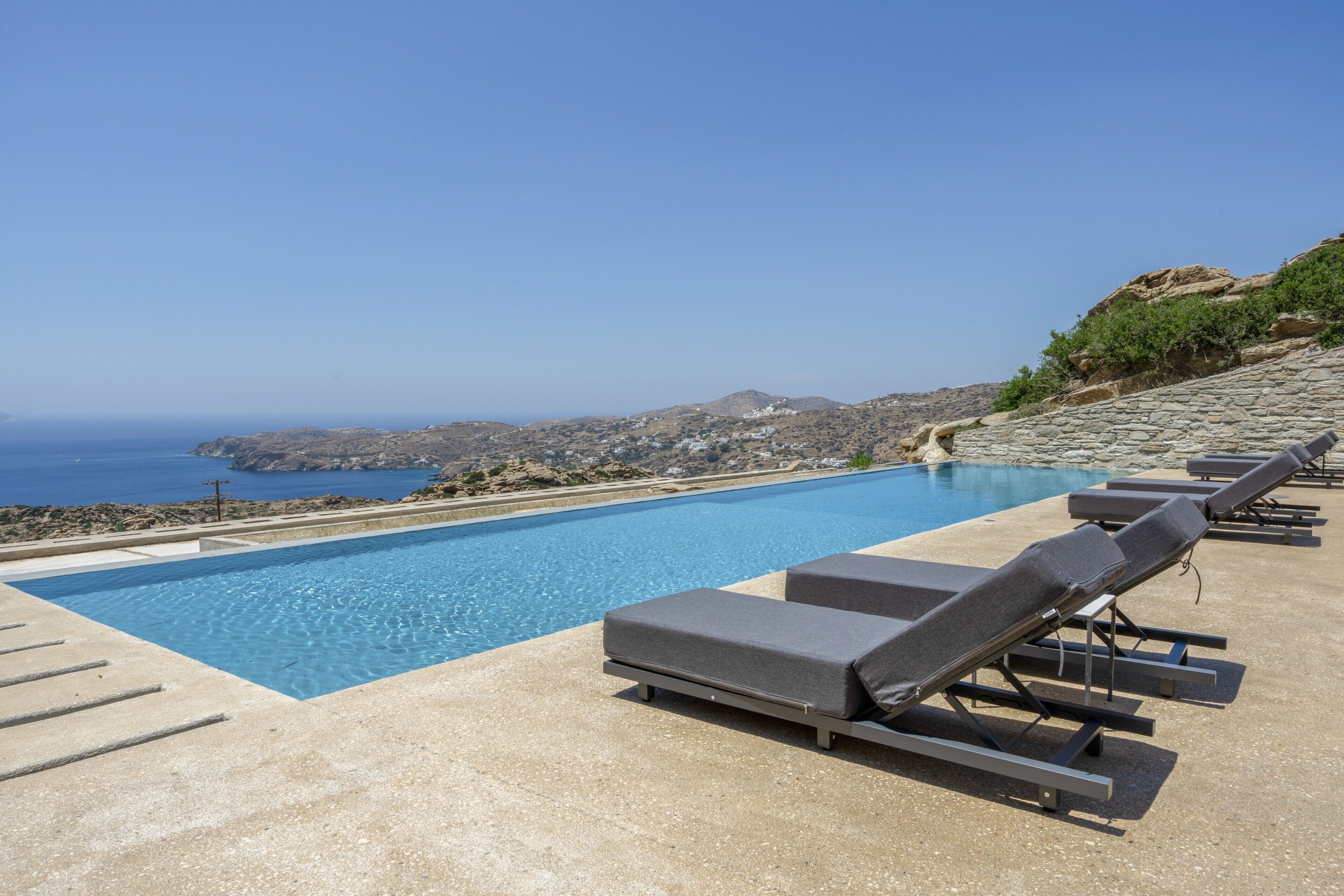 A pool with lounge chairs, a clear sky, and a view of the sea and hills.