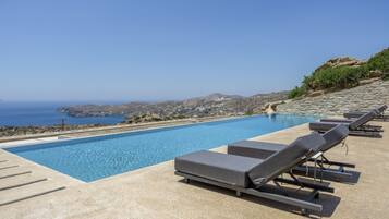 A pool with lounge chairs, a clear sky, and a view of the sea and hills.