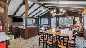 A cozy cabin interior with a wooden dining table, chairs, a fireplace, and a large window.