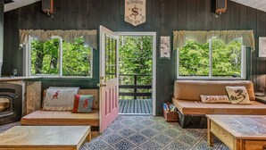 A cozy cabin with a fireplace, two seating areas with cushions, and a large window with a view of trees.