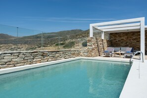 Una piscina con pareti in pietra e un pergolato bianco, circondata da sdraio e vista su delle colline.