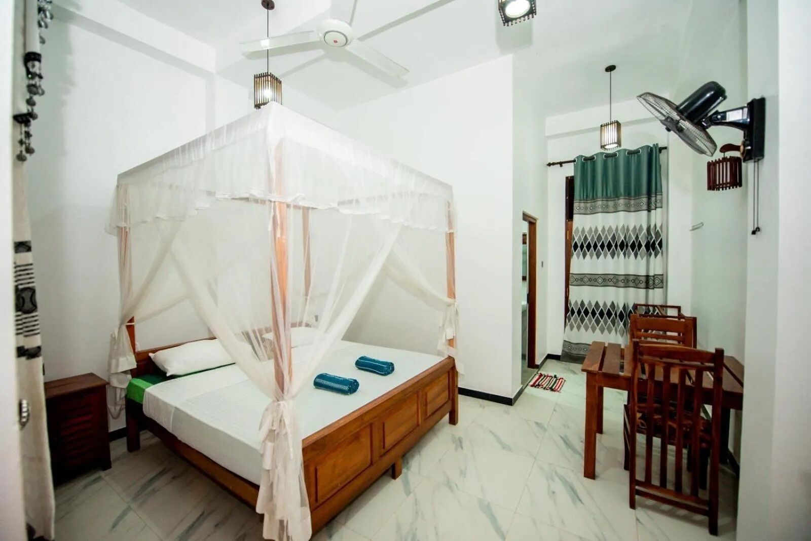 A bedroom with a canopy bed, a ceiling fan, a desk with a chair, and a patterned curtain.