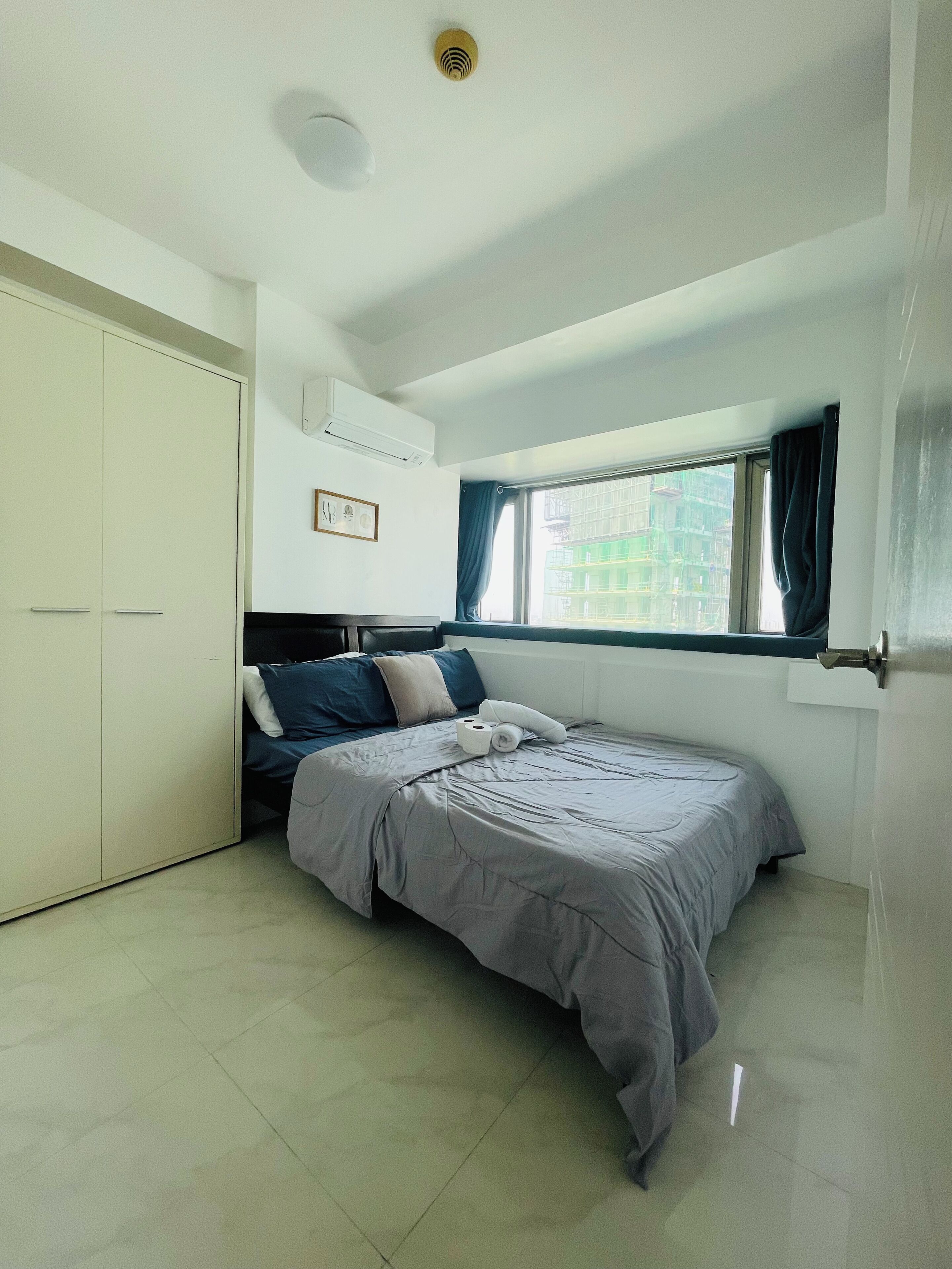 A bedroom with a bed, a wardrobe, an air conditioning unit, a window with curtains, and a view of buildings outside.