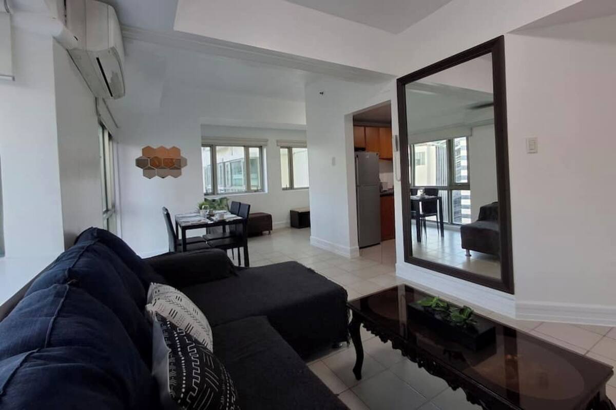 Una sala de estar moderna con un espejo grande, un sofá oscuro, una zona de comedor con mesa y sillas, y una cocina visible a través de un vano abierto.