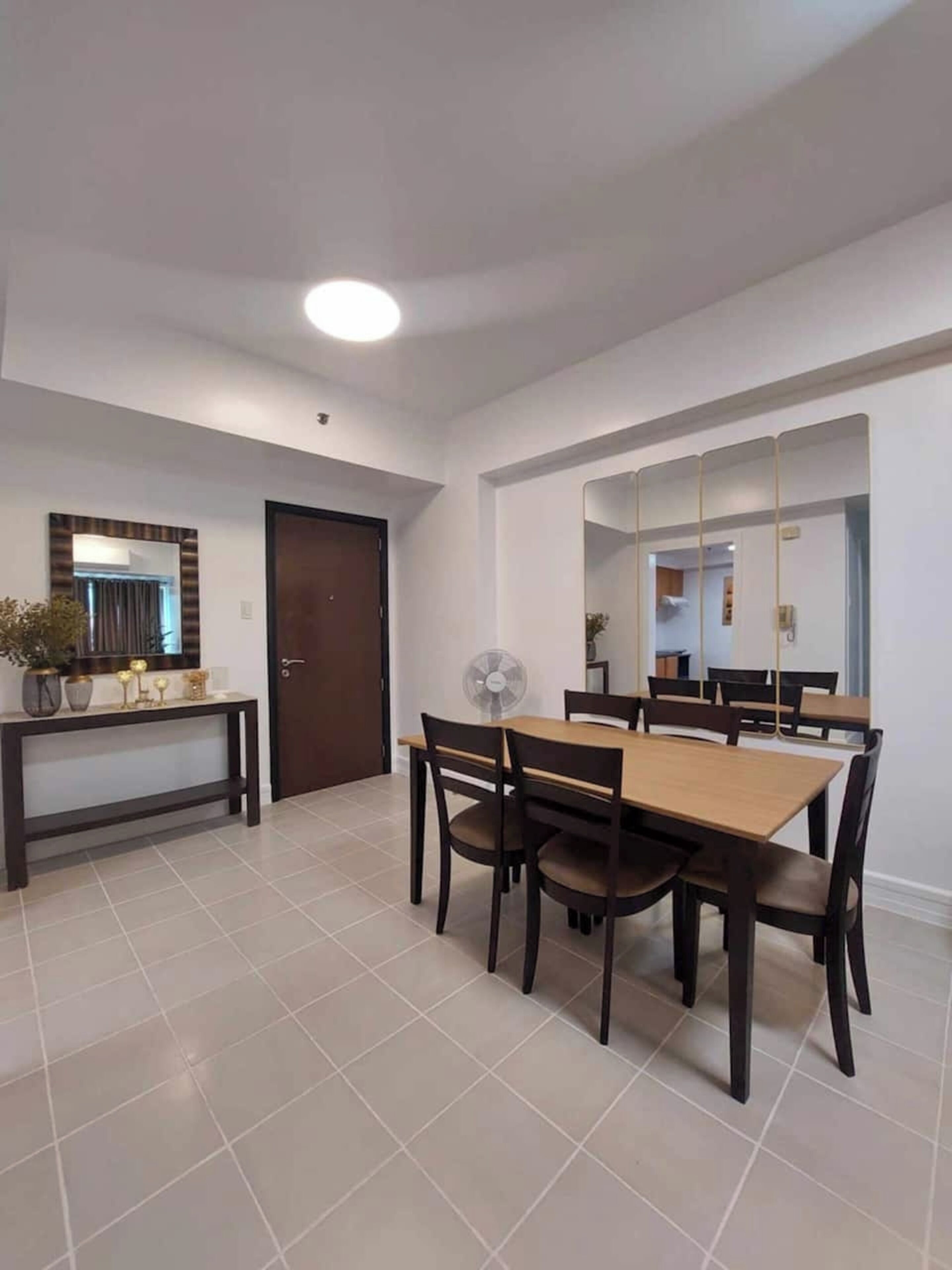 A dining area with a wooden table and chairs, a small console table with decorative items, a door, and a large mirror.