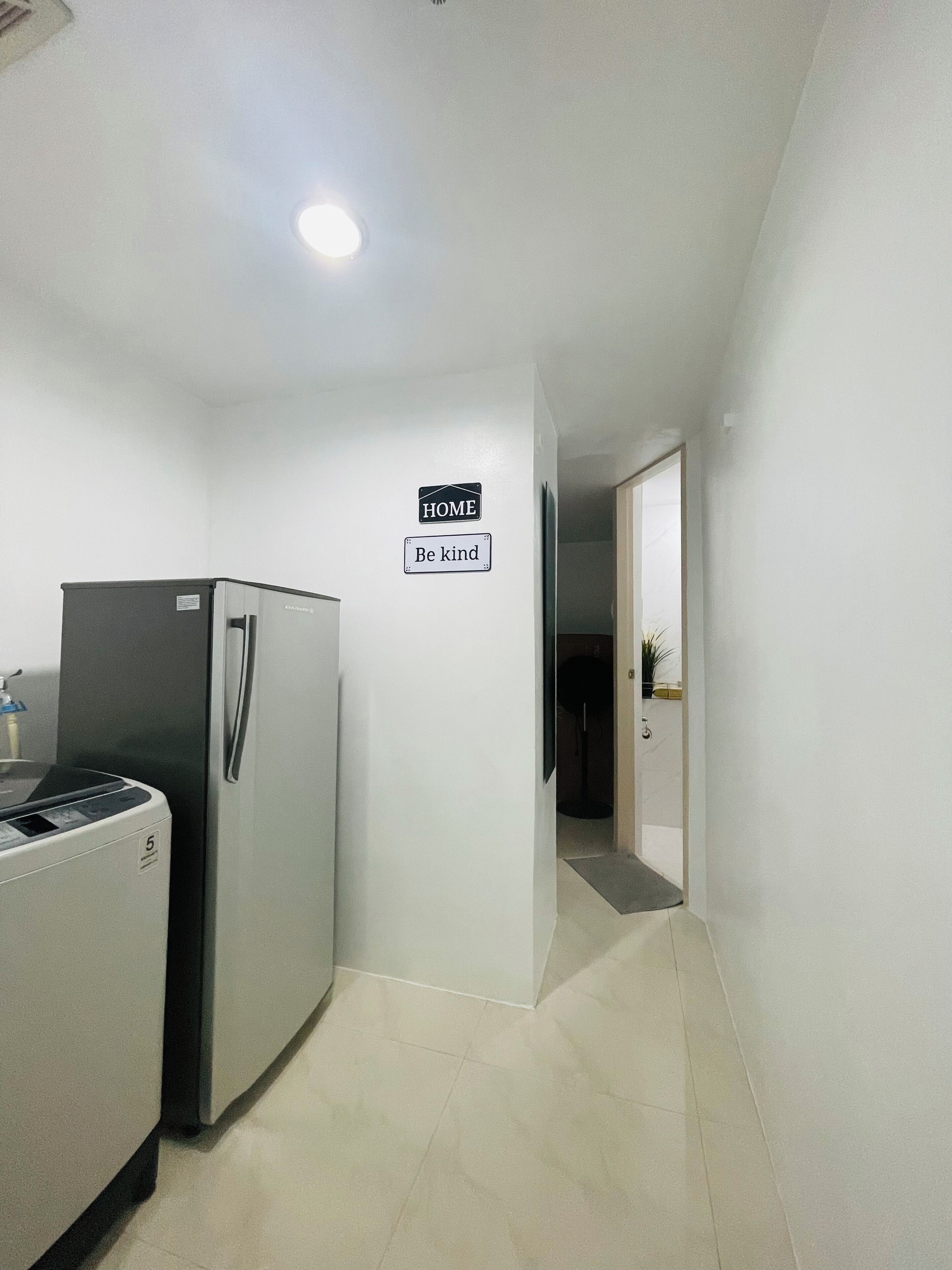 Un couloir avec un réfrigérateur, une pancarte indiquant « DOMICILE » et une autre pancarte sur le mur avec l’inscription « Soyez aimable ».