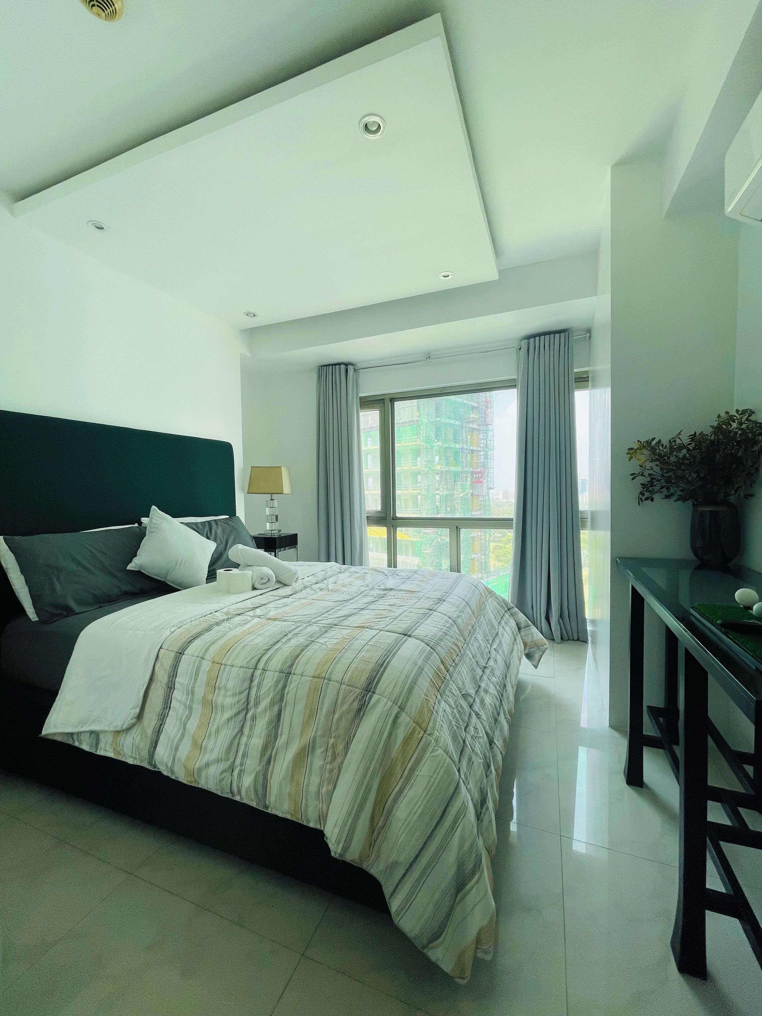A modern bedroom with a large bed, a bedside table, a lamp, and a view of a building through the window.