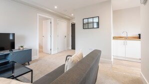 Sala de estar moderna com sofá, TV e uma mesa pequena. Cozinha visível com armários e pia.