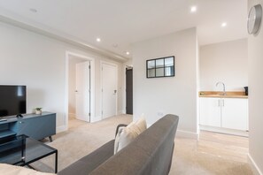 Ein modernes Wohnzimmer mit einer Couch, einem Fernseher und einem kleinen Tisch. Ein Küchenbereich mit Schränken und einer Spüle ist sichtbar.