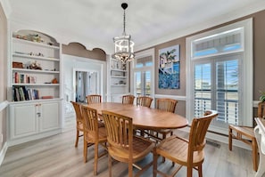 A dining area with a large wooden table, six chairs, a chandelier, and a view through large windows. - Victorian Style Beachfront Home Perfect Family Vacation Destination (Gulf Shores)