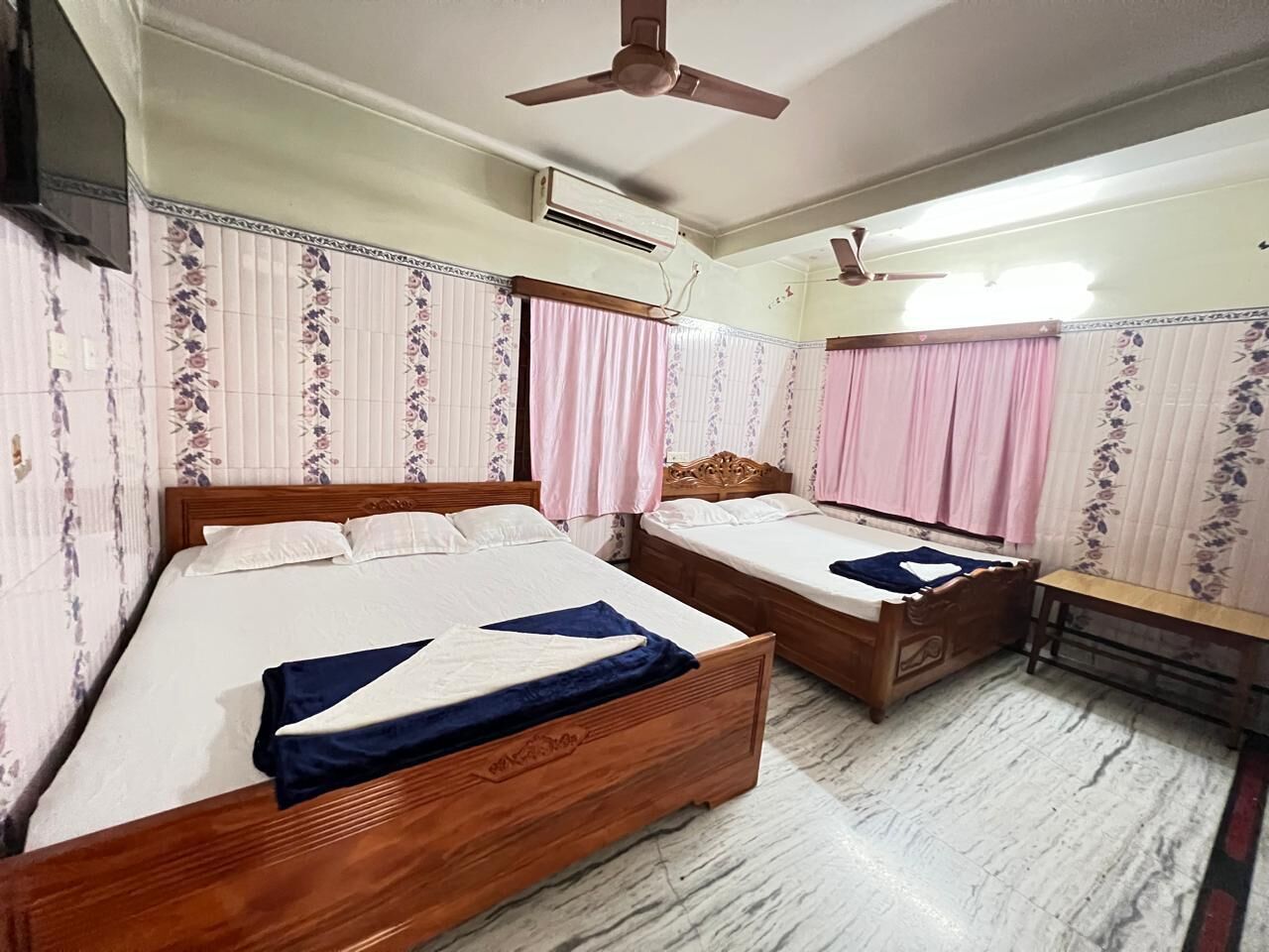 Ein Schlafzimmer mit zwei Holzbetten, einer Bank, einem Deckenventilator, einer Klimaanlage und Wandpapier mit Muster.